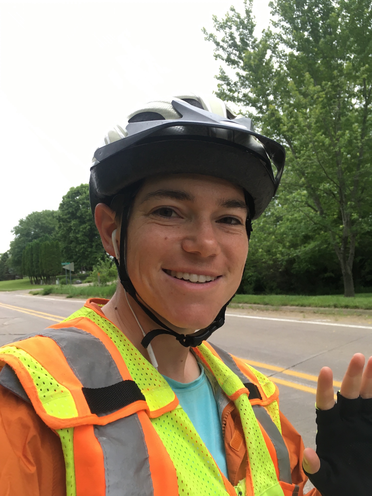 Zeph, equipped with helmet, reflective vest, raincoat, and bike gloves, waves at the camera with trees and a paved road in the background