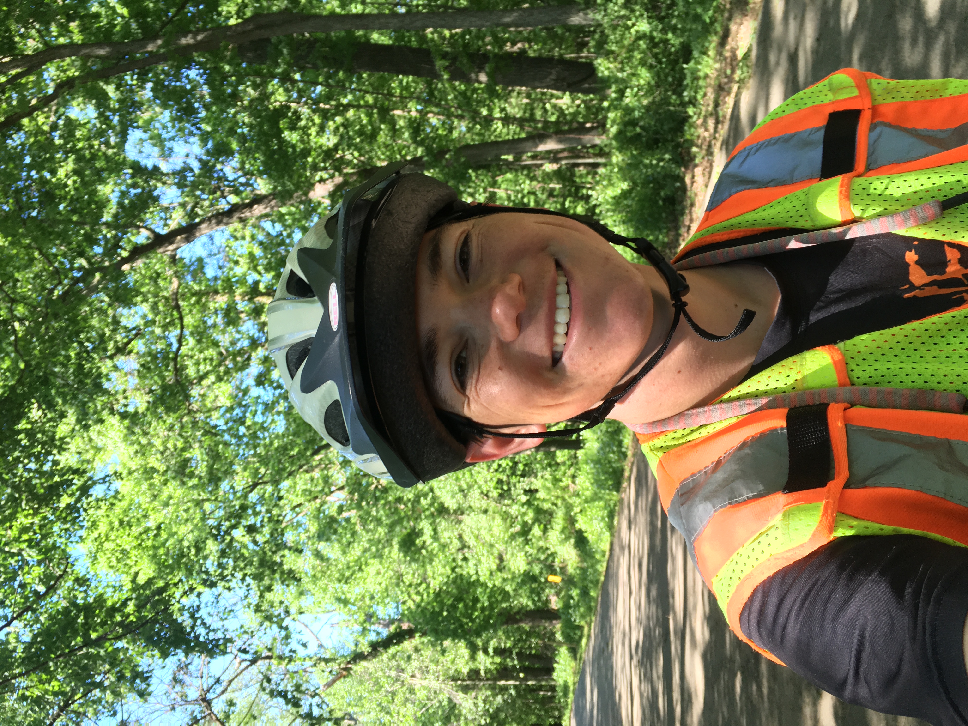 Zeph, equipped with short sleeves, reflective vest and (of course) helmet, smiles at the camera with arching trees overhead creating shade