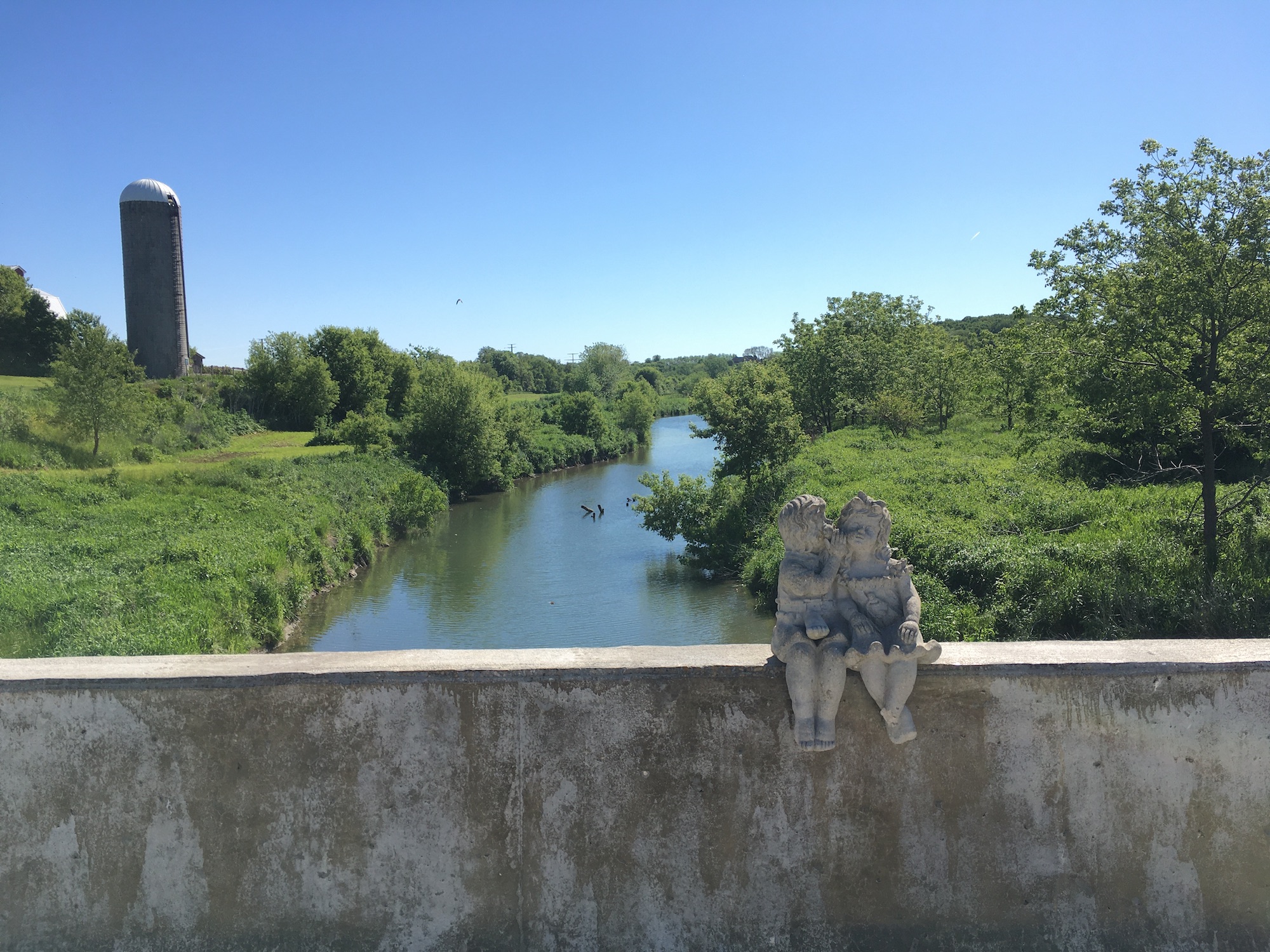 A bridge overlooks a river meandering through green-covered shores including some trees. On the railing of the bridge is a stone statue of two small people sitting side by side. One nuzzles into the other's cheek.