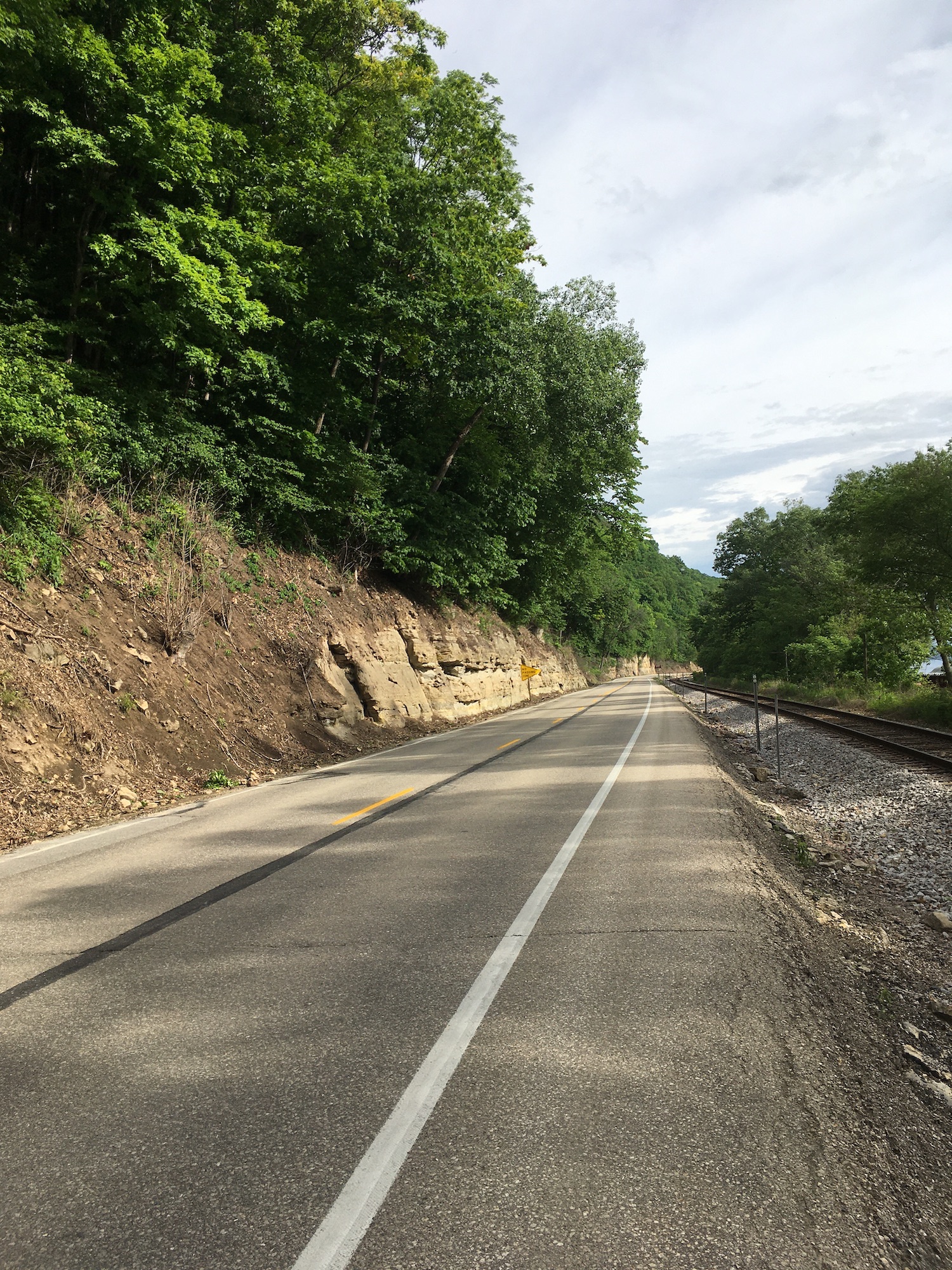 The road vanishes into the distance, with a tree-clad bluff on one side and a sharp drop to a railroad on the other