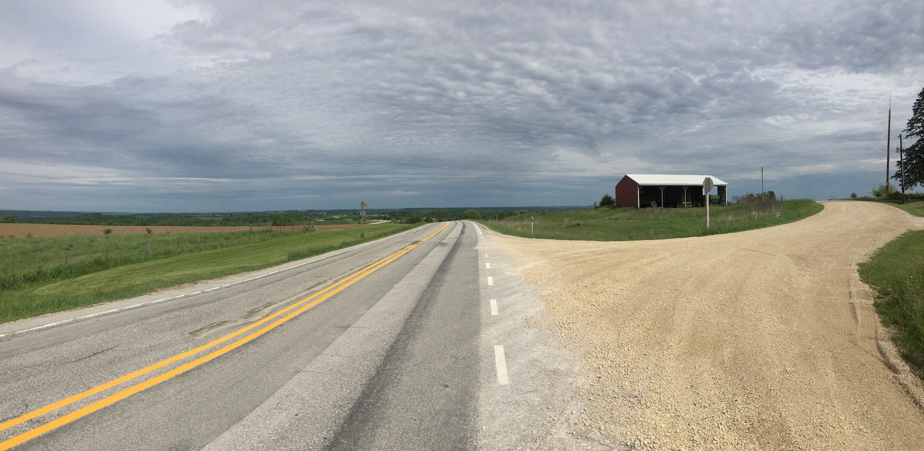 Panorama shot from atop a hill. Rolling hills and farmland/grassland are covered by ripply clouds, and a gravel road leads to a farmhouse on the right