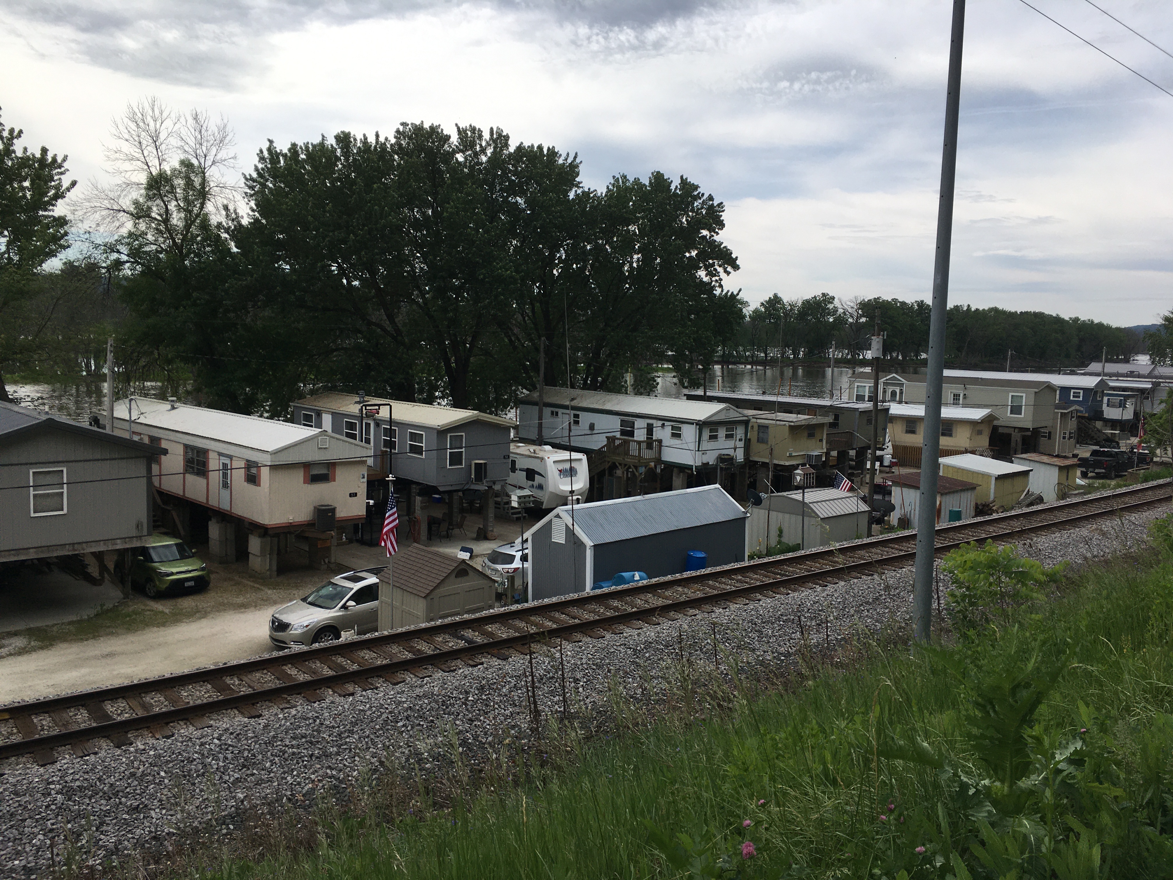 Beyond a railroad, several long, skinny, elevated houses abut the Mississippi River, with trees interspersed.