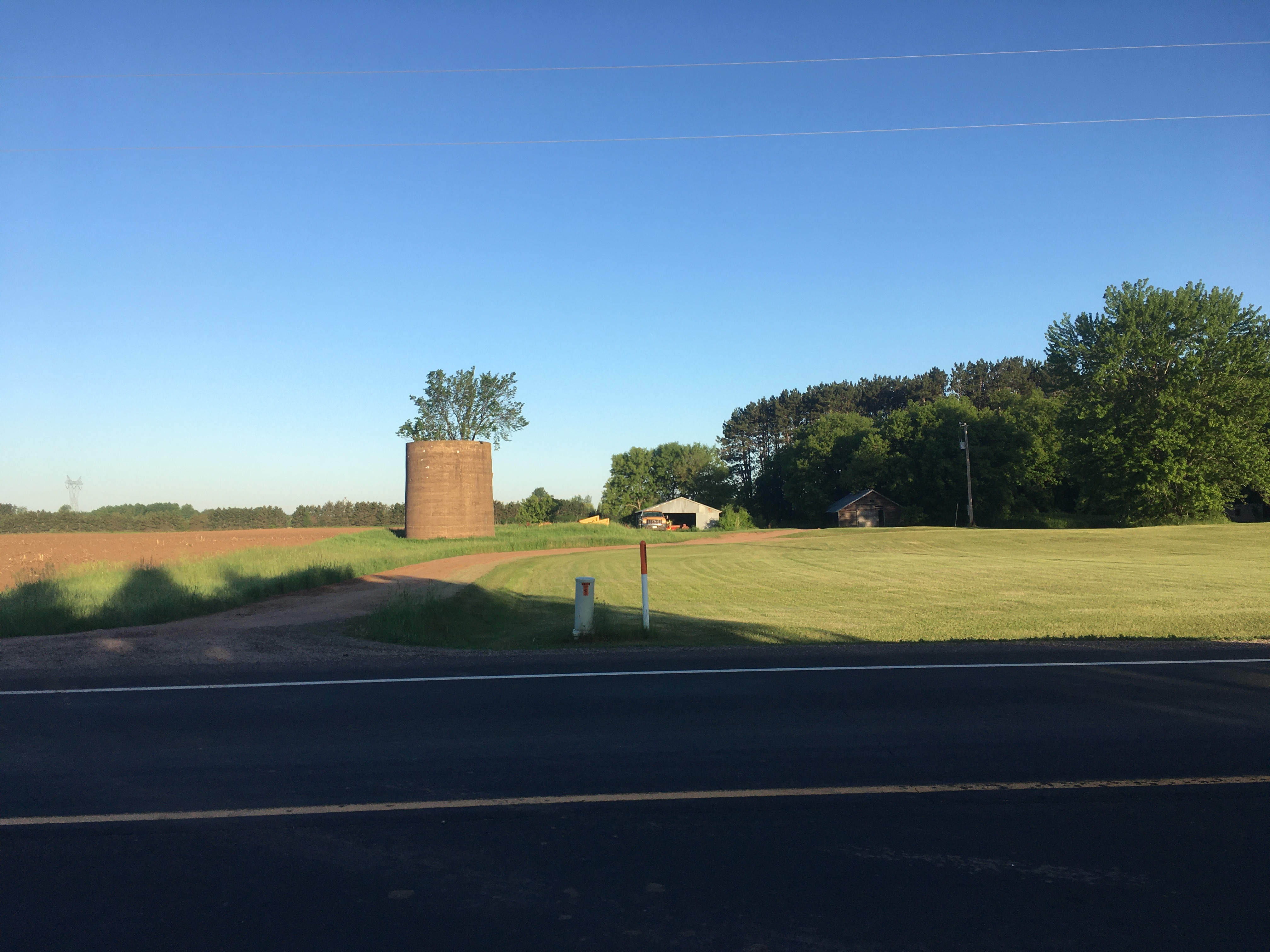 Across a road is a dirt
driveway, and behind it is a cylindrical building, probably 1-2 stories
high, made of concrete or a close relative. It has a tree growing out of
the middle of it. Power lines, a small home, and a denser patch of trees
are also visible.