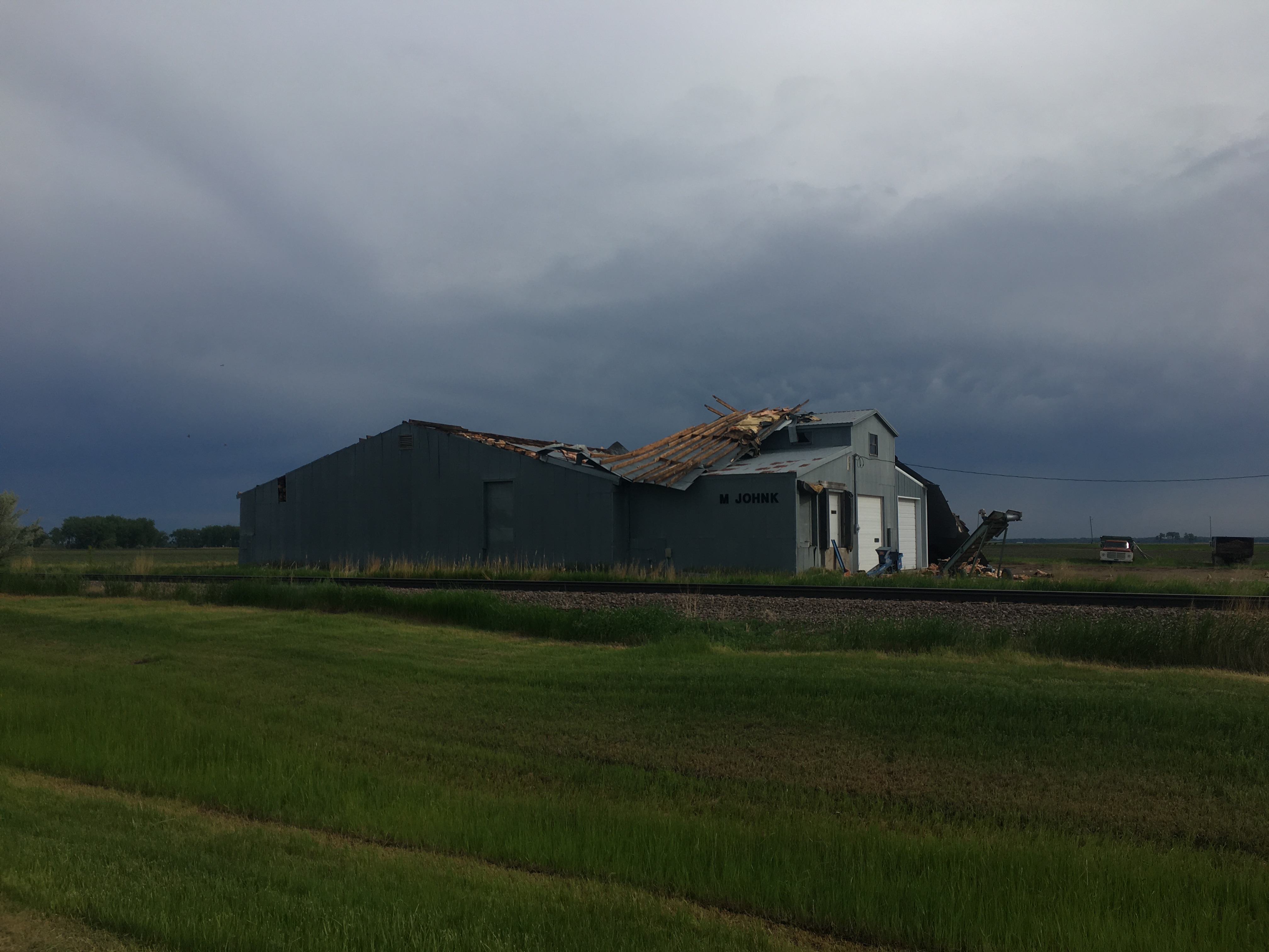 A large single-story wooden
building in a field is shown, with half its roof ripped off and lying
upside down from the wind. Ominous dark gray clouds in the background.