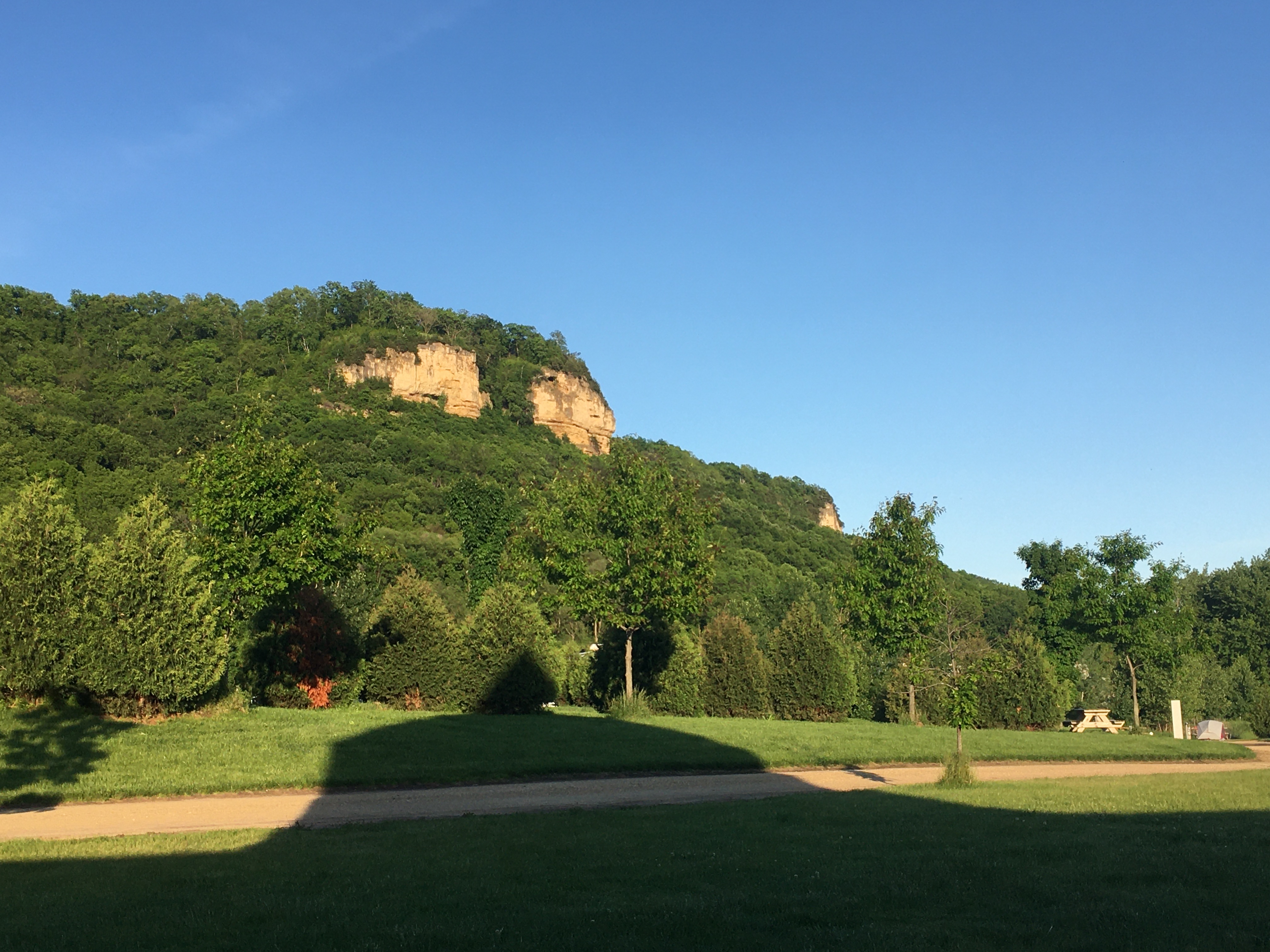 Gorgeous view from my campsite – a bluff rises up from grassy and forested flat areas. A shadow stretches upward towards the bluff. In the distance, a small tent (mine) can be seen.