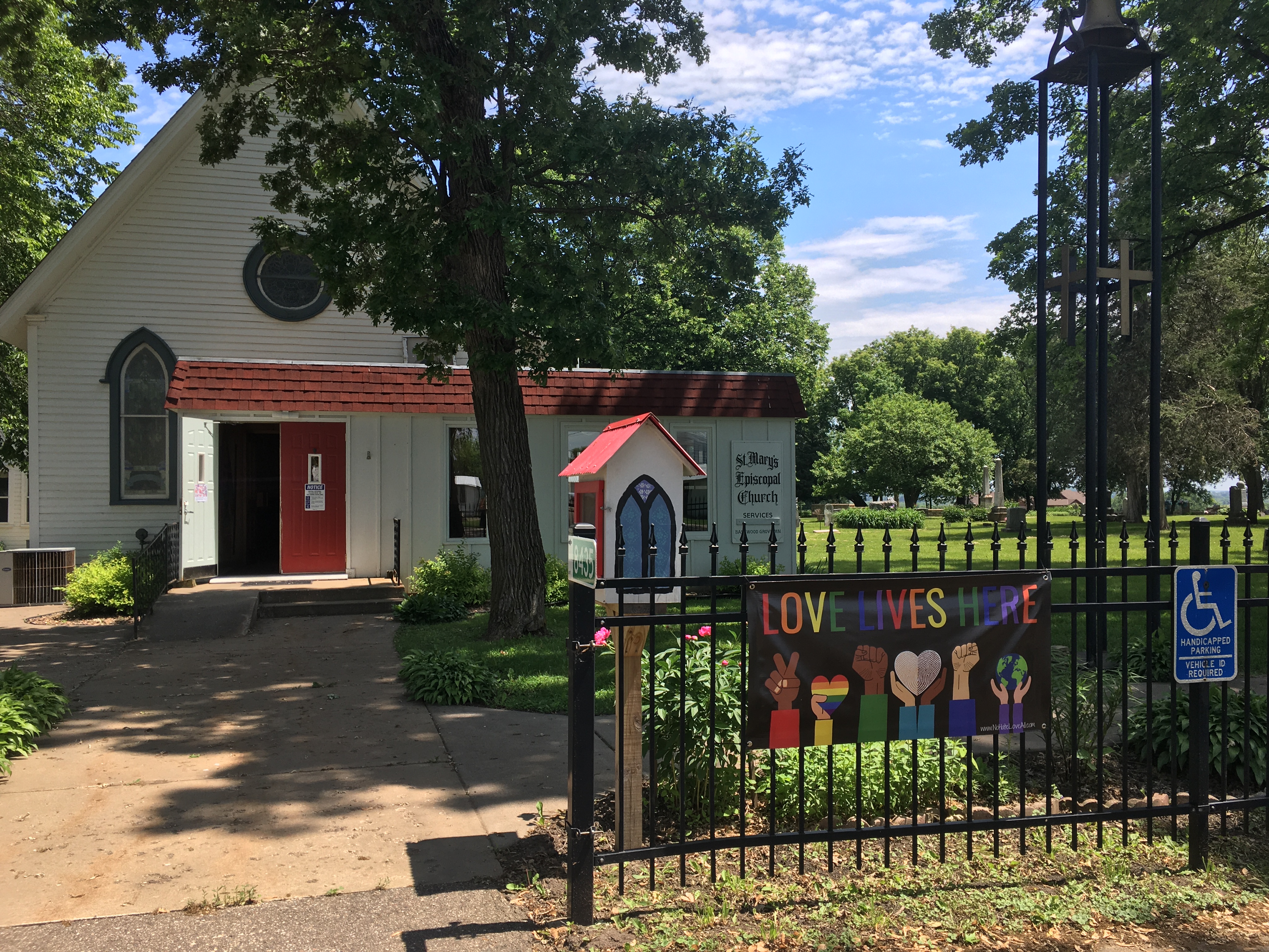 A decorative fence in front of a small church building. The fence has a large sign saying LOVE LIVES HERE with a variety of inclusive symbols, including diverse skin colors, a rainbow heart, a black power fist, an interracial marriage heart-shaped fingerprint, and a globe.