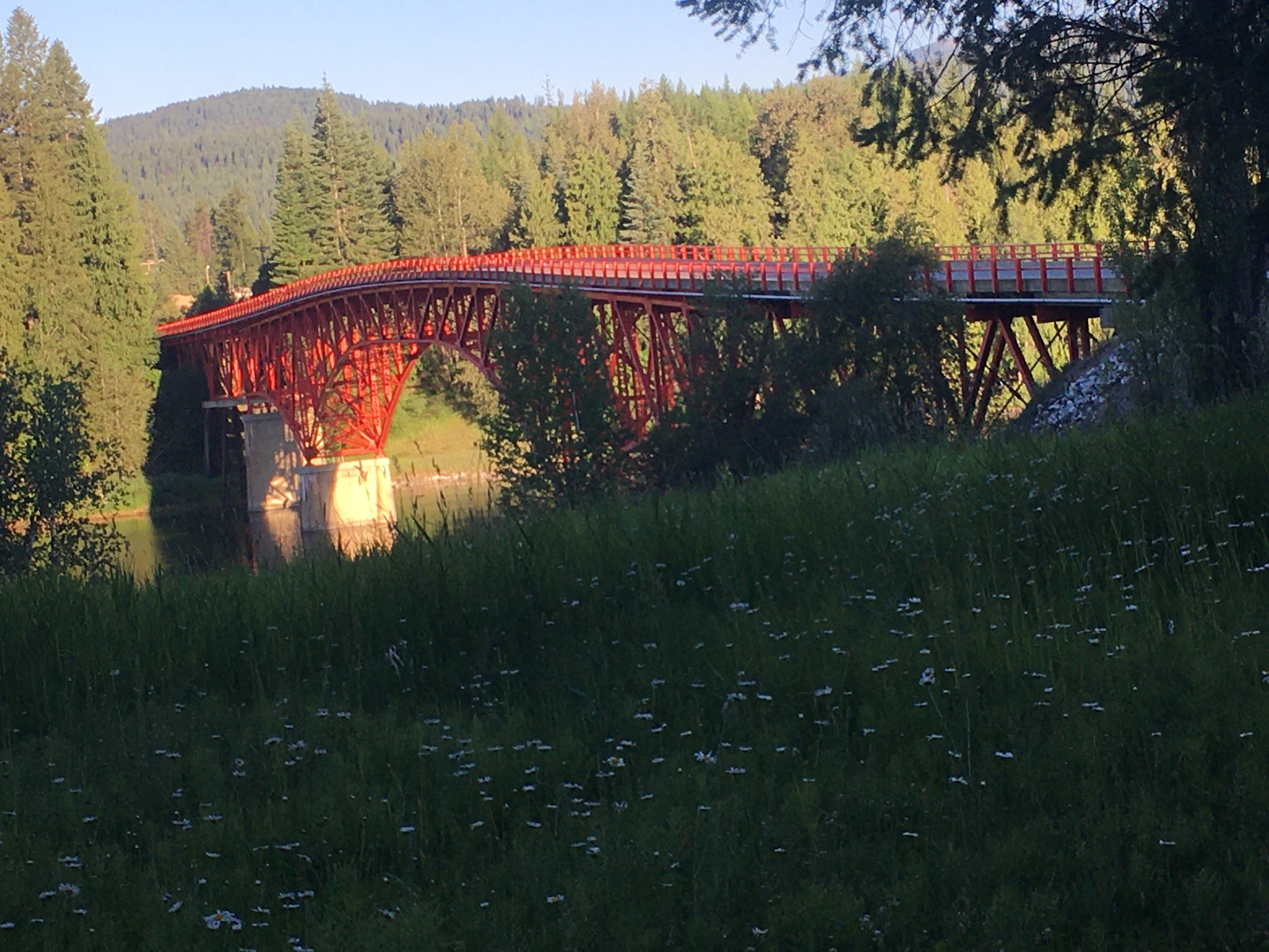 A red bridge spans a wide
river, with grass in the foreground and treed hills behind